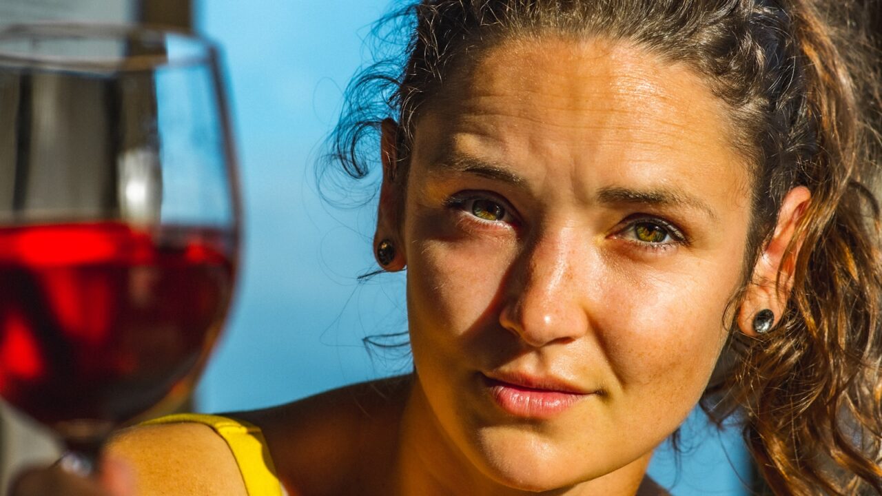 Een vrouw met krullend haar en oorbellen houdt een glas rode wijn dicht bij haar gezicht en kijkt recht in de camera. Ze draagt een geel topje en wordt verlicht door zonlicht. De achtergrond is onscherp.