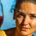 Een vrouw met krullend haar en oorbellen houdt een glas rode wijn dicht bij haar gezicht en kijkt recht in de camera. Ze draagt een geel topje en wordt verlicht door zonlicht. De achtergrond is onscherp.