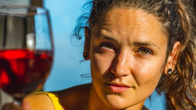 Een vrouw met krullend haar en oorbellen houdt een glas rode wijn dicht bij haar gezicht en kijkt recht in de camera. Ze draagt een geel topje en wordt verlicht door zonlicht. De achtergrond is onscherp.
