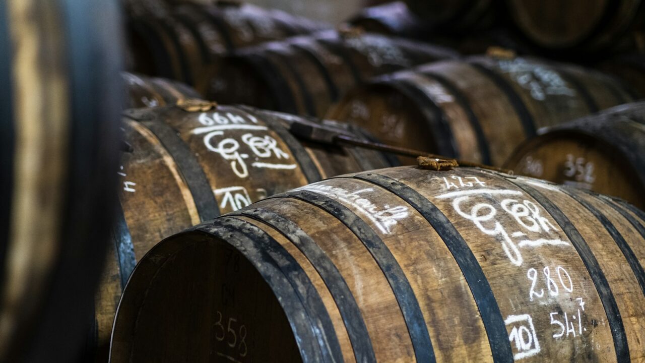 Verschillende houten vaten met metalen banden staan opgestapeld in een opslagruimte. Witte krijtmarkeringen met cijfers en letters staan op de uiteinden van de vaten. De scène is schemerig verlicht.